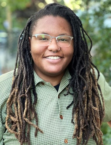 A portrait of a Black woman with long hair, glasses, and a green shirt.